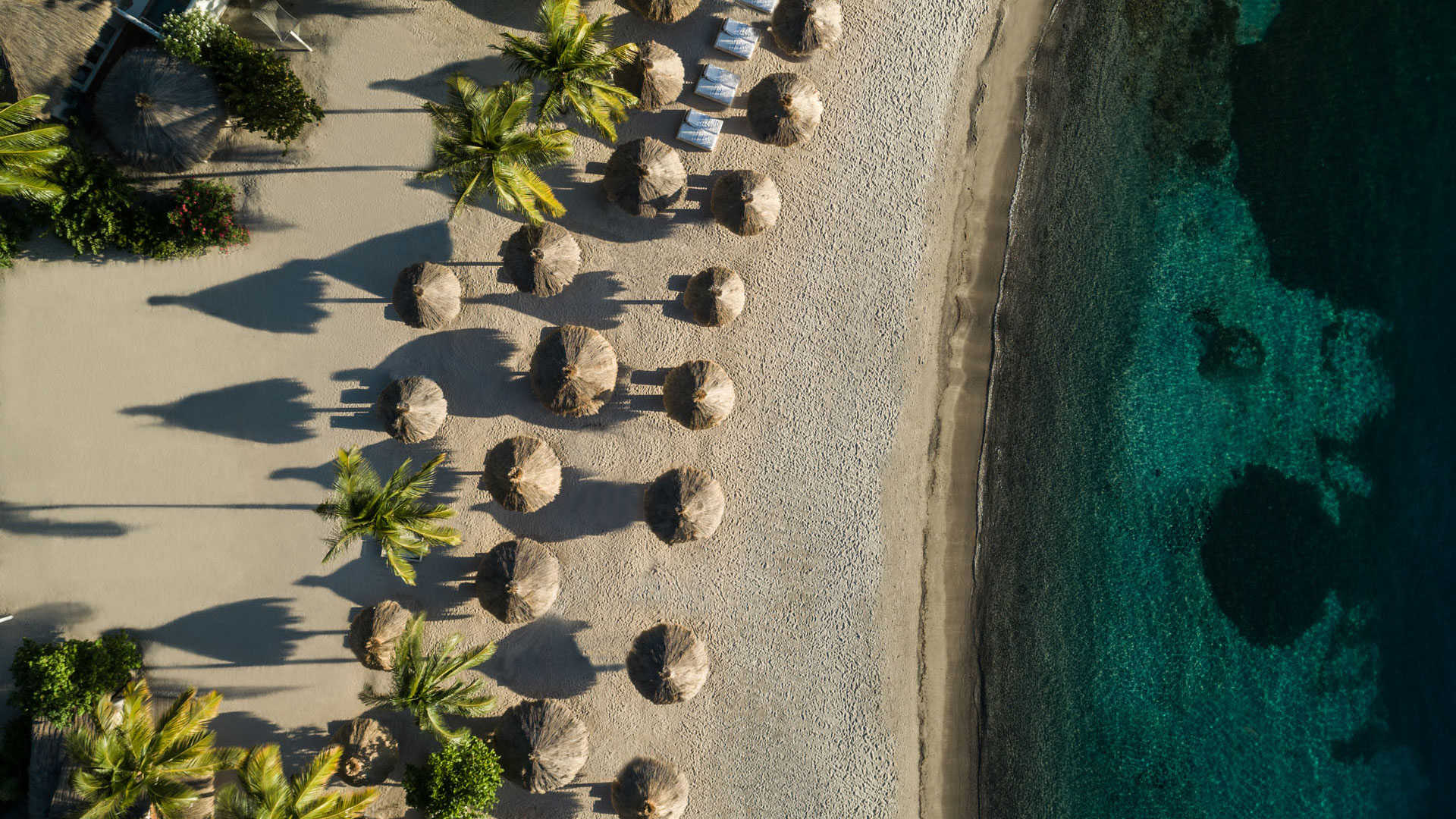 Aerial view of a sandy tropical beach with palm trees, thatched umbrellas, lounge chairs, and clear turquoise water.