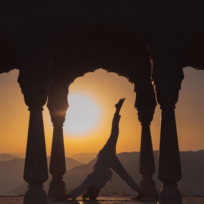 Silhouette of a person doing yoga at sunset beneath an ornate pavilion, with mountains in the distance.
