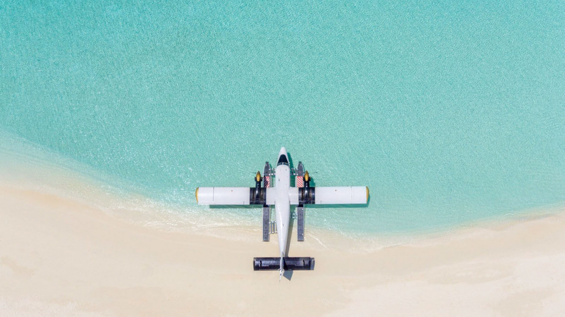 Aerial view of a seaplane parked in shallow turquoise water beside a white sand beach.