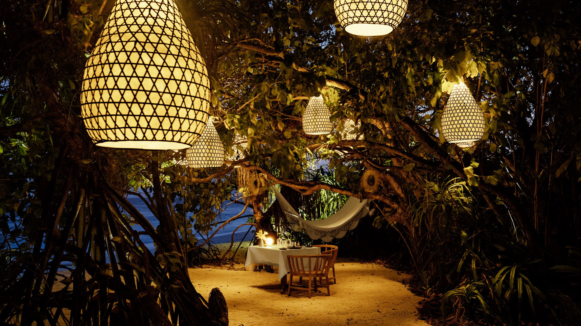 Romantic outdoor dining setup under glowing lanterns in a lush tropical grove at night.