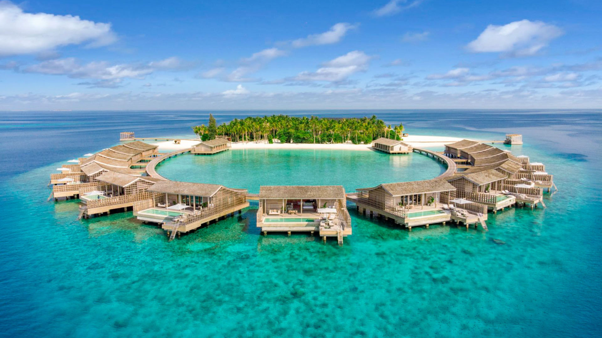 Circular overwater villas surround a lagoon beside a small tropical island in clear blue sea.