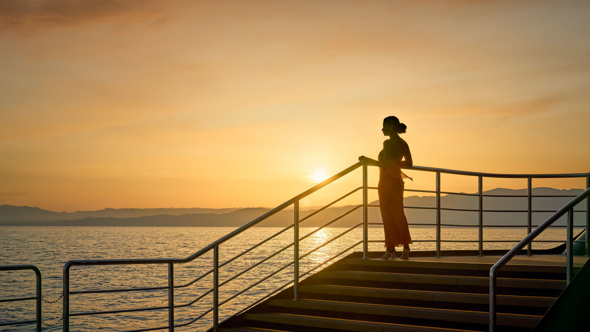 Silhouette of a woman standing on deck aboard The Ritz-Carlton Yacht Collection at sunset, overlooking calm sea and distant mountains.