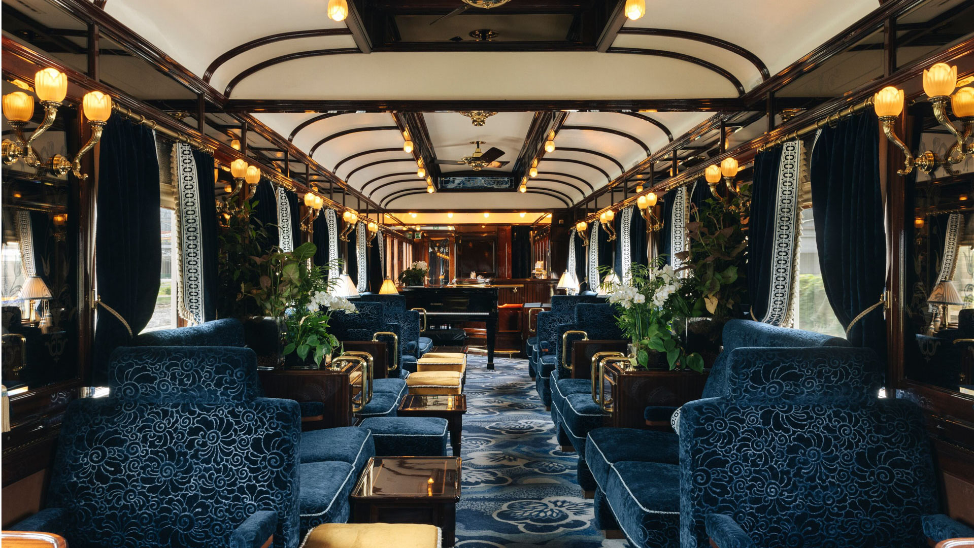 Opulent lounge carriage on the Venice Simplon-Orient-Express with plush blue armchairs, polished wood details, soft lighting, and a piano at the far end.