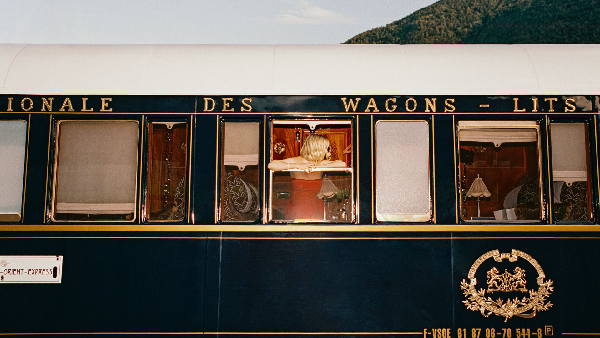 Passenger leaning out of a window on the blue-and-gold Venice Simplon-Orient-Express carriage, with mountain scenery in the background.