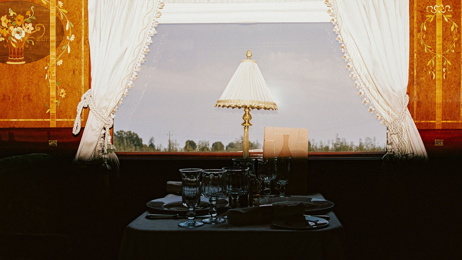 Elegant dining table beside a curtained window inside the Venice Simplon-Orient-Express, with ornate wood marquetry and a lamp framing the countryside view.