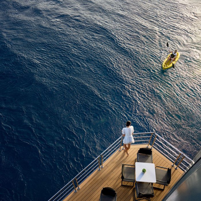 Guest standing on a private terrace aboard The Ritz-Carlton Yacht Collection, looking out over the sea as a kayaker paddles nearby.