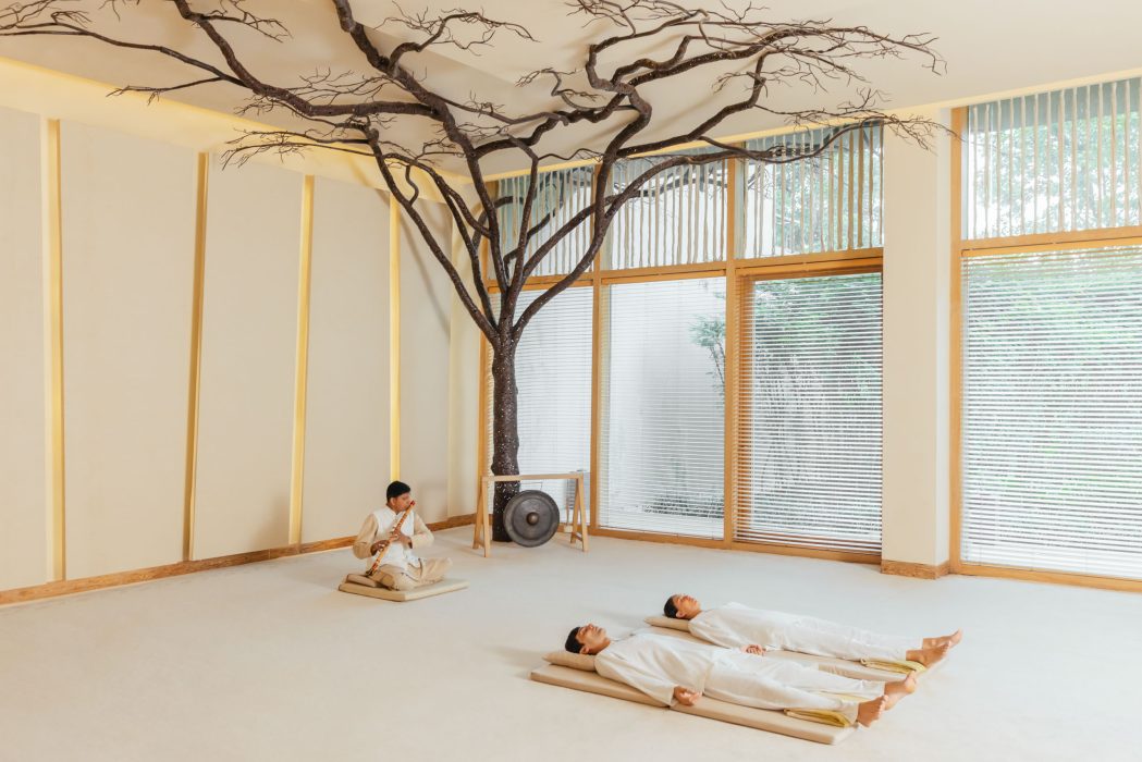 Two guests lie on floor mats during a meditative sound therapy session at Six Senses Vana, while a musician plays flute beneath a sculptural tree installation.