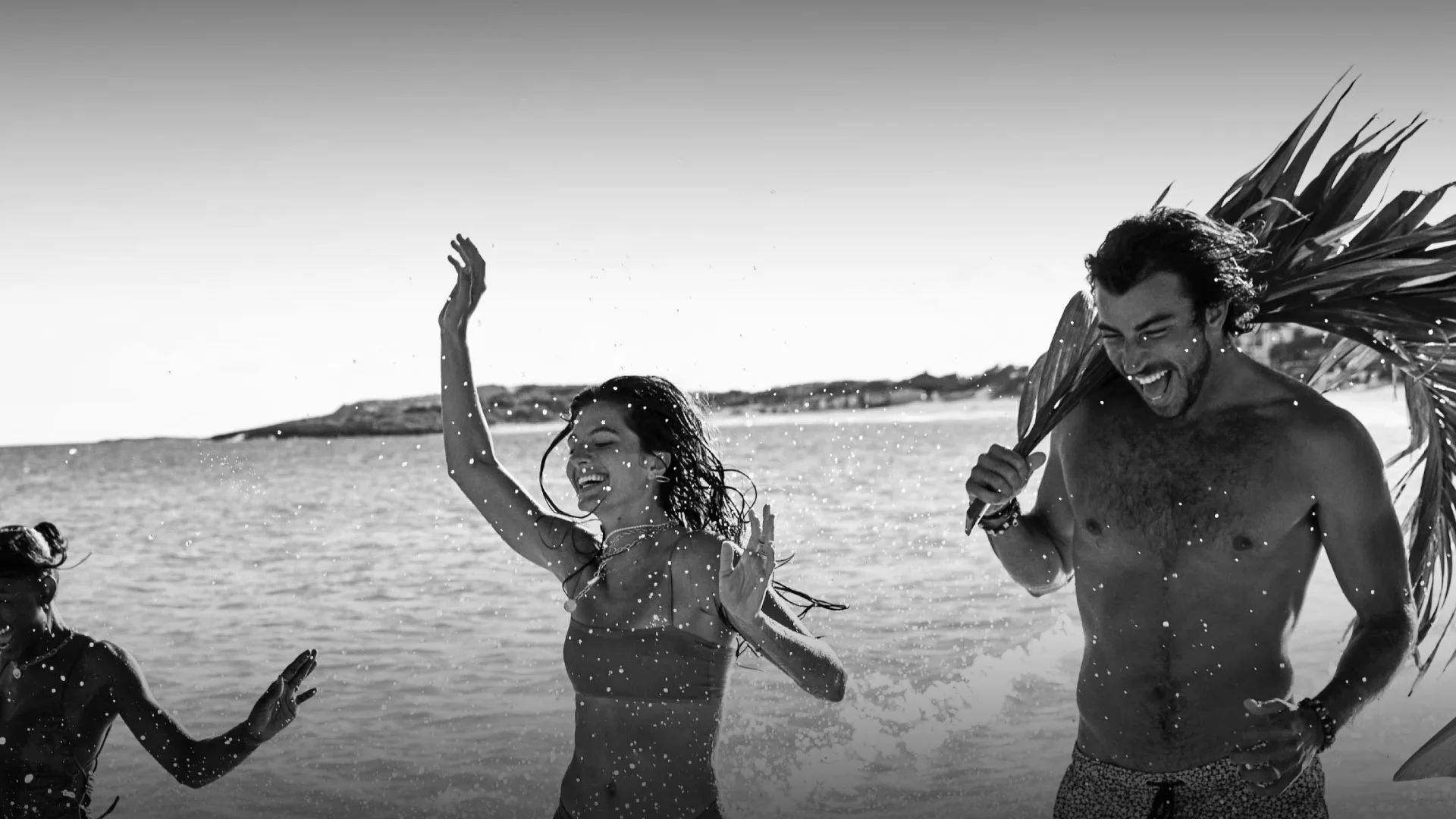 Adults laugh and splash in the shallow sea at Belmond Cap Juluca beach, one holding palm fronds overhead.