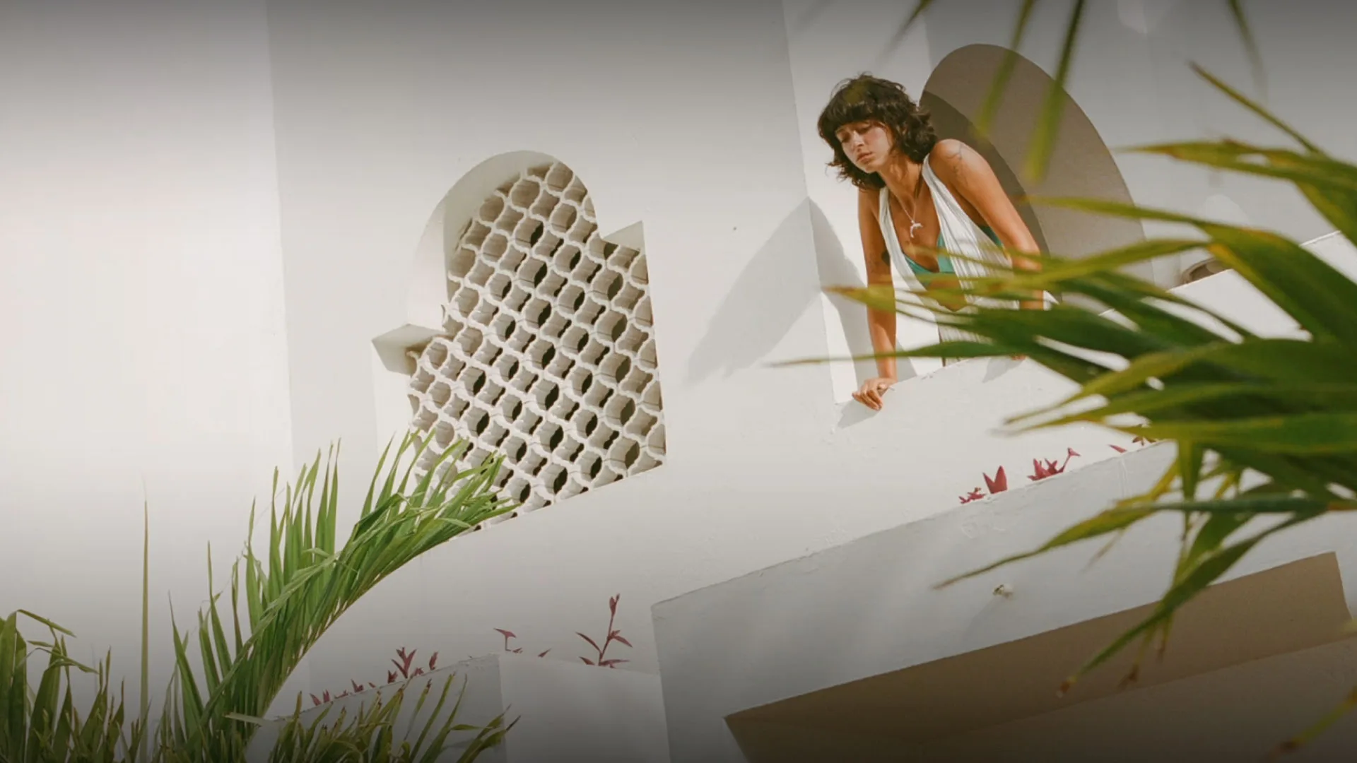 Woman leaning over a white stucco balcony at Belmond Cap Juluca, framed by tropical plants and arched architectural details.