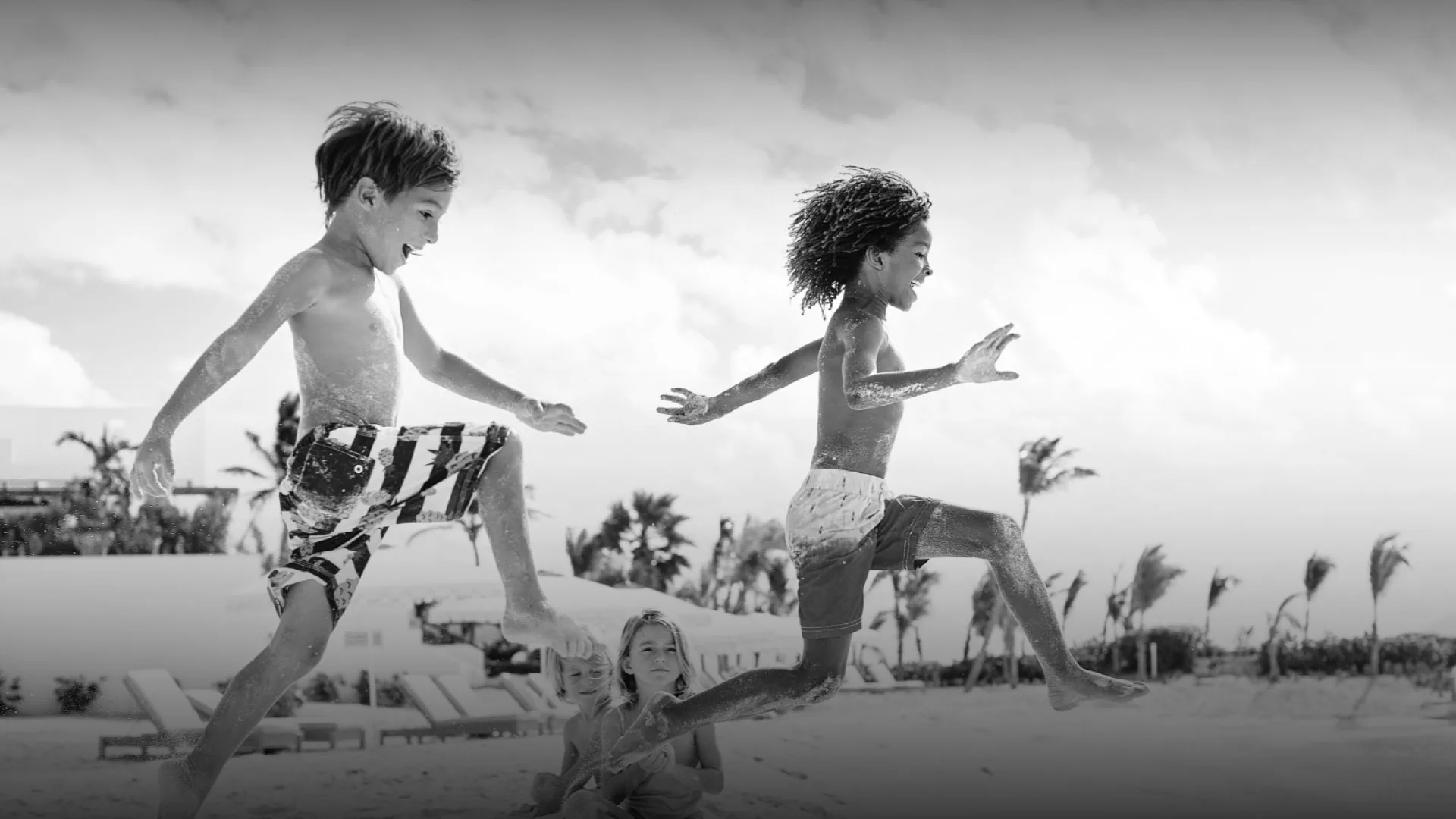 Children play and jump on the sand at Belmond Cap Juluca, with palm trees and beachfront loungers in the background.