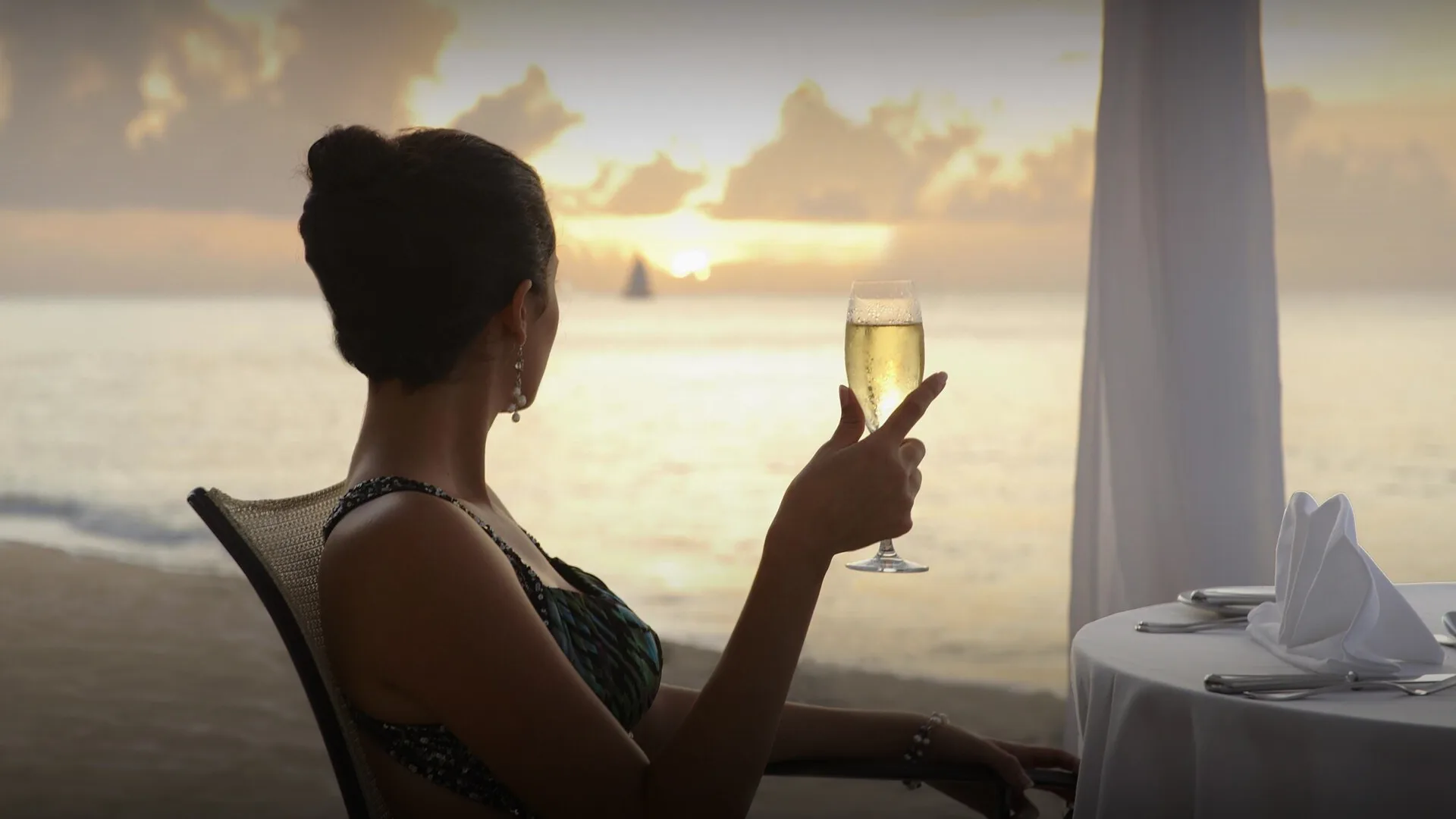 Woman holding a glass of champagne while watching the sunset from a beachfront table at Fairmont Royal Pavilion, Barbados.