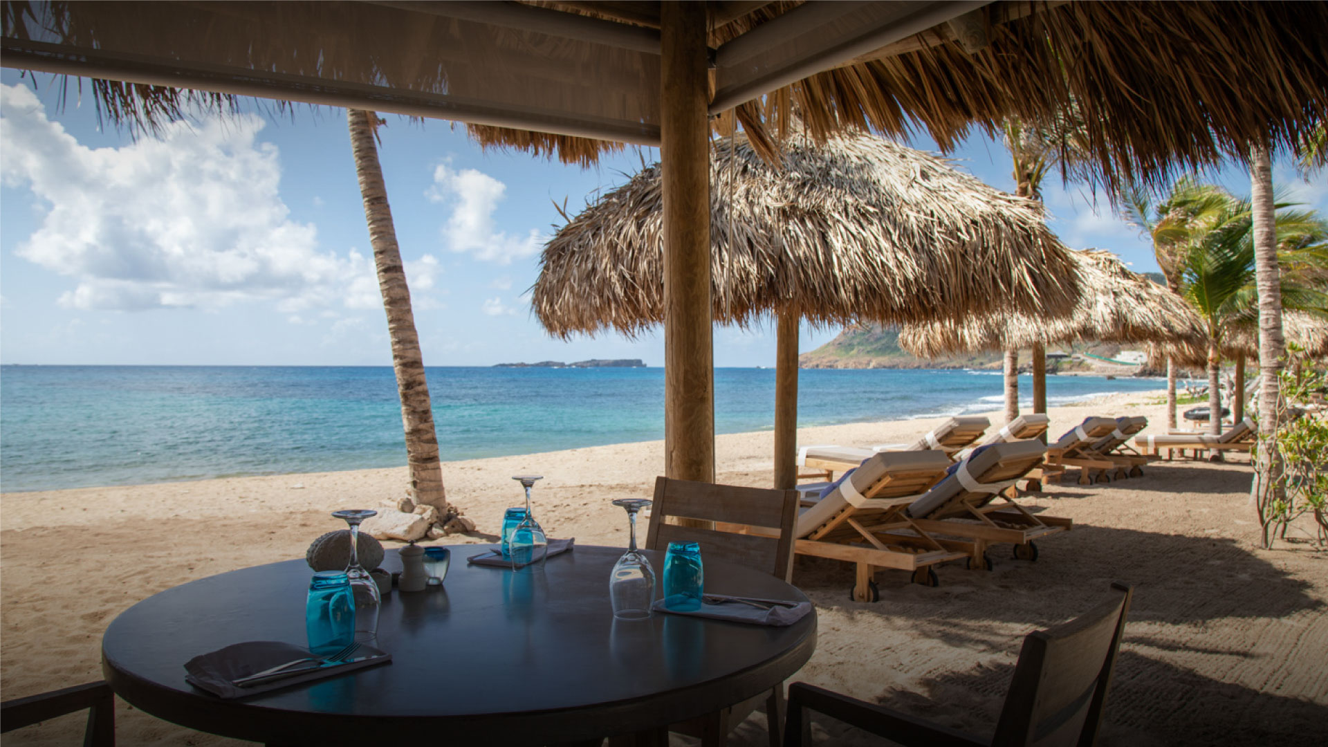 Beachfront dining table under thatched roof with lounge chairs and ocean view at Hotel Le Toiny
