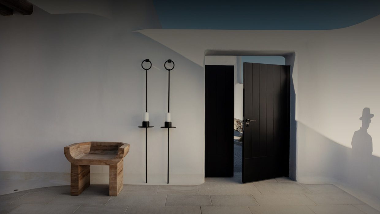 Minimalist whitewashed entrance courtyard with black doors and carved wooden chair at Kalesma Mykonos