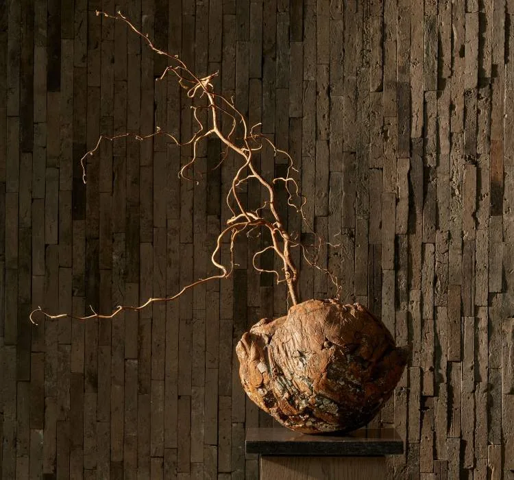 A sculptural root and bare branch on a plinth against a wall of stacked timber