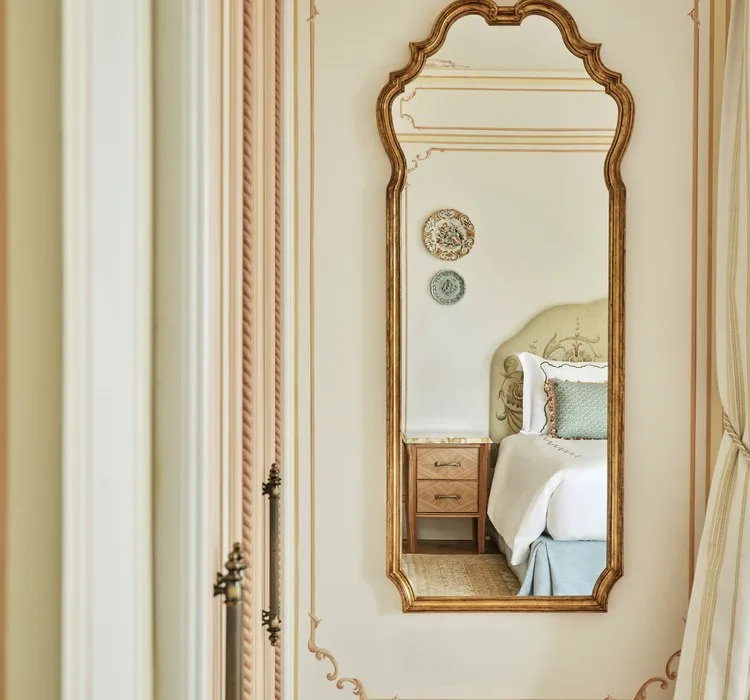 A gilt antique mirror reflecting a corner of a bedroom, headboard and nightstand, beside hand-painted wall panelling