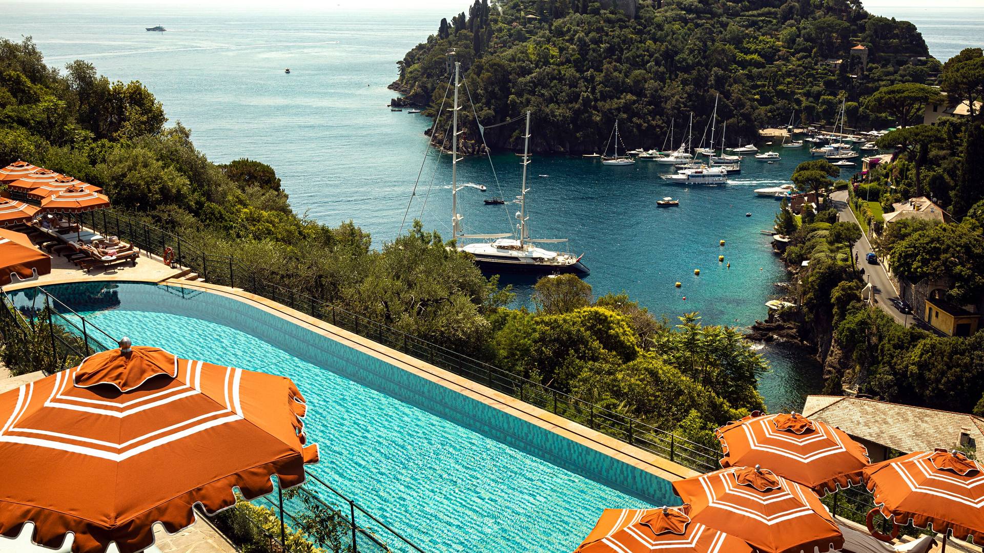 The hotel's infinity pool and orange-striped umbrellas overlooking Portofino harbour, yachts moored below a wooded headland