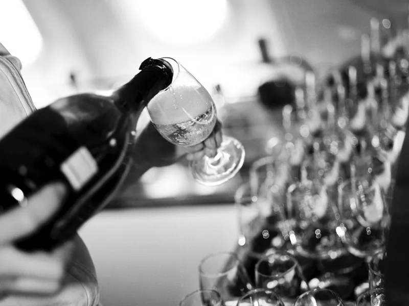 Champagne poured into a glass beside a long row of empty glasses on board, in black and white