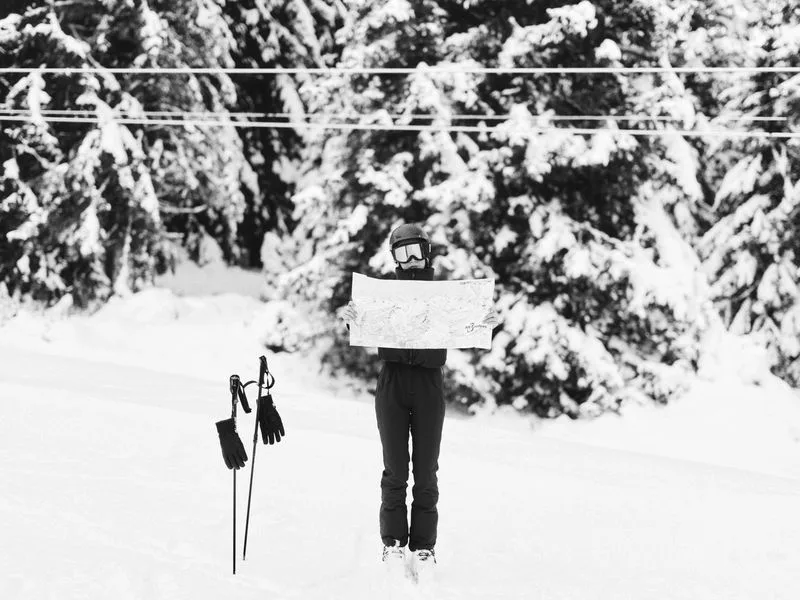 A skier holding up a piste map, face hidden behind it, in black and white