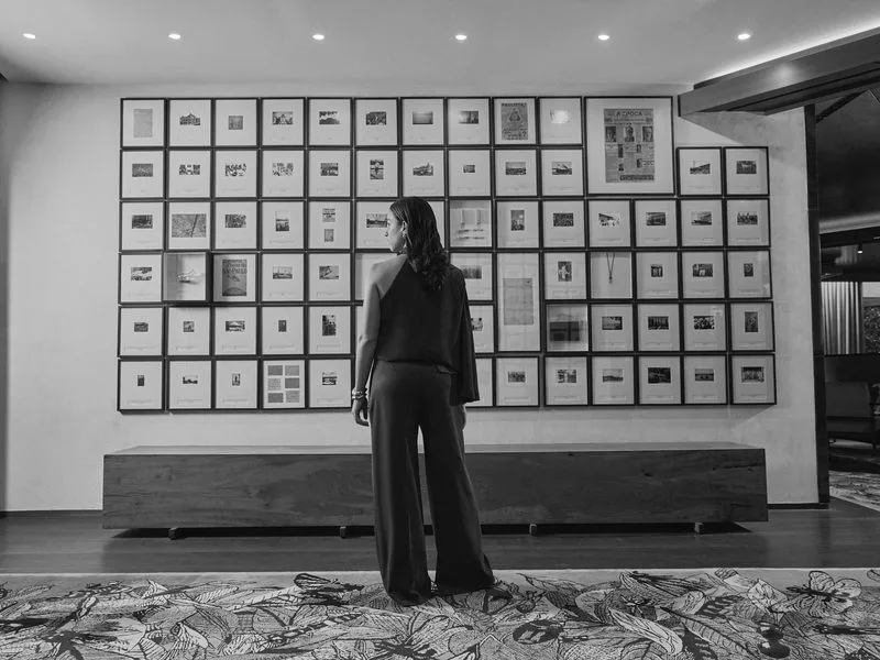 Woman in a jumpsuit viewed from behind, facing a gallery wall of framed archival photographs and ephemera, mono