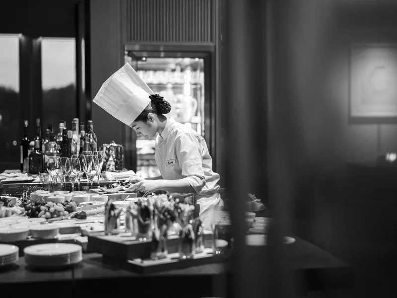 A chef in whites at work behind a counter of glassware and fruit, in black and white