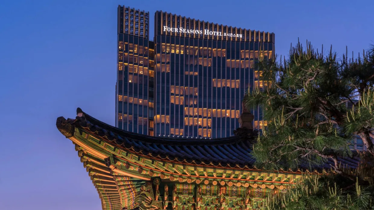 The tower of Four Seasons Hotel Seoul rising behind a painted palace eave and a pine at dusk