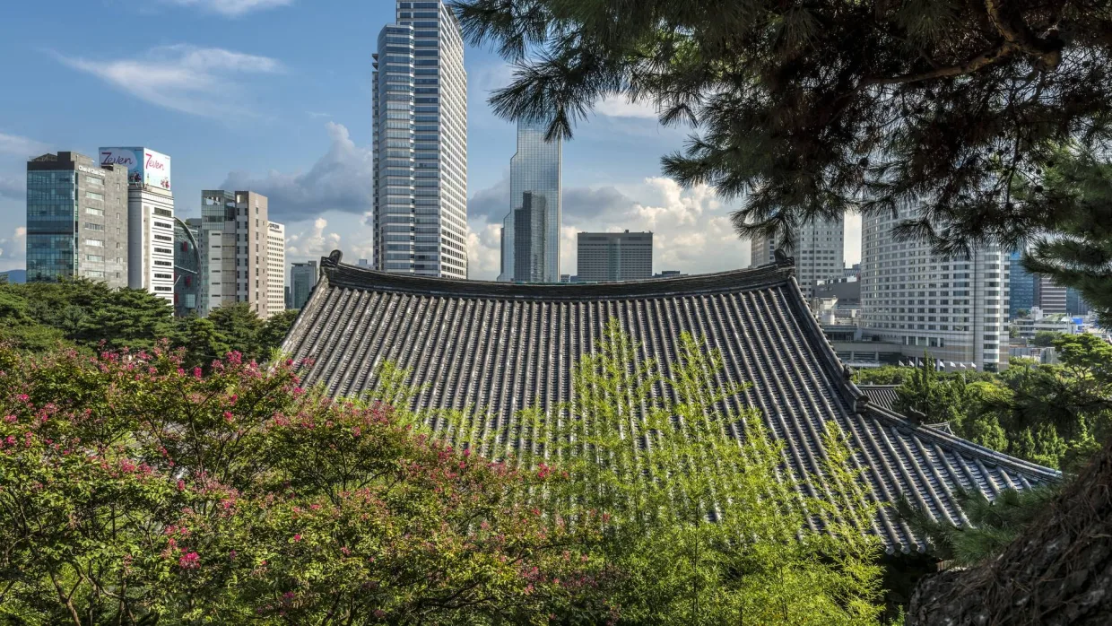 A tiled hanok roof among summer trees with the Seoul skyline behind