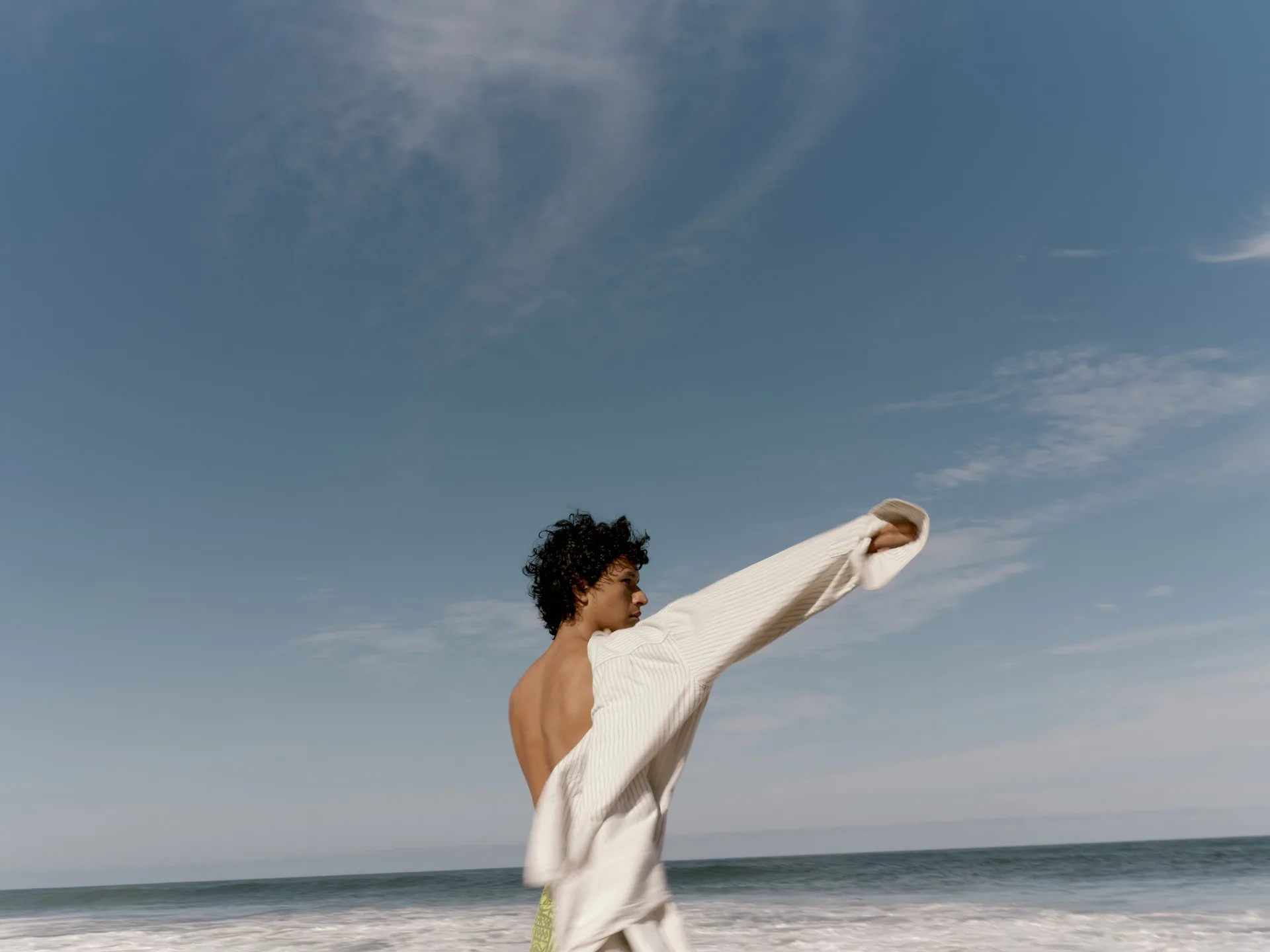 A guest on the beach at Rosewood Mandarina, a loose white shirt caught in the wind against a blue sky