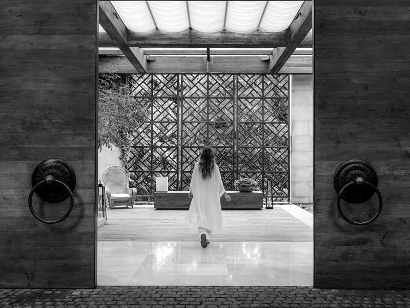 Black-and-white view through tall timber doors into the open lobby pavilion at Rosewood Phuket, a guest walking towards a lattice screen and garden